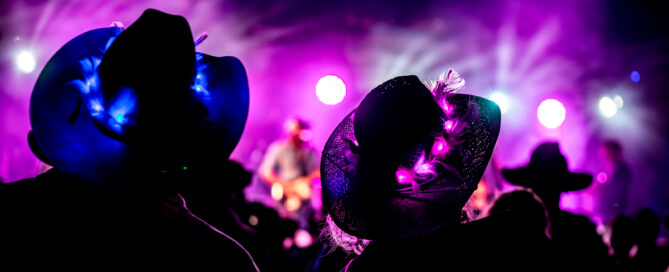 Silhouetted concert crowd wearing wide-brim hats, with a guitarist on a purple-lit stage in the background.
