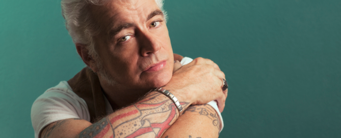 Portrait of a gray-haired man with tattoos resting his arms on a teal studio backdrop, looking at the camera with a serious expression.