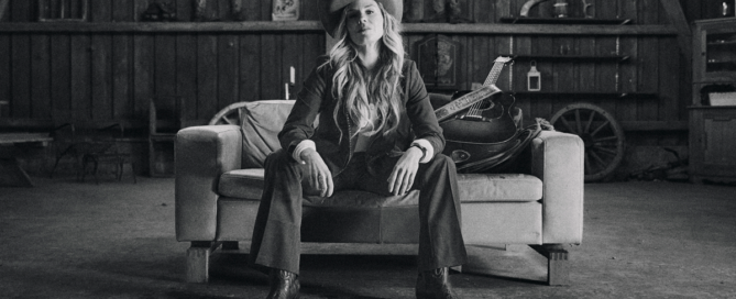 Woman in a cowboy hat sits on a sofa in a rustic barn, with a guitar resting beside her on the couch.